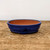 A side-angle view of a shallow, oval blue glazed bonsai pot resting on a rustic wooden surface. The exterior of the ceramic container is finished in a vibrant, deep cobalt blue glaze with a glossy sheen and subtle variations in color, giving it a handmade feel. A distinctive horizontal ridge wraps around the center of the pot's exterior for added texture. 

The interior remains unglazed, showing the warm, natural terracotta-brown color of the clay. The pot features small, integrated feet at the base to allow for proper drainage and stability. It is positioned on a weathered wooden table characterized by visible wood grains and surface scratches, set against a minimalist off-white background that highlights the pot's elegant form and striking blue color.