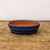 A high-resolution product photograph shows a shallow, oval-shaped ceramic bonsai pot sitting in the center of a rustic wooden surface. The pot features a deep, mottled blue glazed exterior with subtle variations in tone, giving it a textured and artisanal look. The interior of the pot remains unglazed, revealing the natural, warm reddish-brown color of the terracotta clay.

The pot’s design includes a smooth, rounded top rim and a single decorative horizontal ridge that wraps around the upper third of its body, adding a touch of classical elegance. It is slightly elevated by small, integrated feet at the base, which allow for proper drainage and airflow. The pot is placed on a weathered, light-brown wooden tabletop that shows natural grain and some signs of wear, all set against a plain, off-white background that makes the rich blue color of the glaze stand out. This professional-grade glazed bonsai pot is designed to provide a beautiful and functional home for a variety of bonsai tree species.