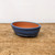 An oval-shaped ceramic bonsai pot sits centered on a rustic wooden surface against a plain, off-white background. The pot features a deep, textured navy blue glaze on its exterior, while the interior is left unglazed, showing the natural reddish-brown color of the terracotta clay. The pot has a smooth, rounded rim and is elevated by subtle, integrated feet. The weathered wooden tabletop below has a light brown tone with visible grain, scratches, and markings, providing a grounded, organic feel to the composition. The soft, even lighting highlights the speckled details in the blue glaze and the smooth, professional design of the bonsai container.