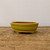 A shallow, oval glazed ceramic bonsai pot in a vibrant mustard yellow finish sits on a rustic wooden surface against a plain, off-white background. The pot features a smooth, glossy exterior with a pronounced top rim and a secondary decorative horizontal ridge encircling the upper portion. The interior of the pot is unglazed, showing the natural reddish-brown clay body. It is supported by small, integrated, curved feet that provide a slight elevation. The bright yellow color and classic oval shape make it a standout choice for various bonsai styles.