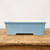 A front-facing, eye-level shot of a rectangular, light blue glazed ceramic bonsai pot. The pot has a soft, matte finish in a pale sky blue color, with a thick, rounded rim around the top edge. Its design features a slightly tapered body that leads down to a base with blocky feet, created by a wide, rectangular cutout in the center of the long side. The interior of the pot is a contrasting creamy off-white. The container is resting on a weathered, rustic wooden surface with visible grain and texture, set against a solid, neutral white background.