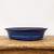 A side-profile photograph of a shallow, oval-shaped glazed bonsai pot in a vibrant shade of cobalt blue. The ceramic pot features a slightly flared rim and a decorative horizontal band near its base, resting on several small, integrated feet. The deep blue glaze has a subtle, mottled texture, while the unglazed reddish-brown clay is visible along the very bottom edge. The pot sits centered on a rustic, weathered wooden board with a prominent grain and visible wear. The entire scene is set against a clean, neutral off-white background, highlighting the rich color and elegant form of the bonsai container.