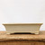A front-facing, eye-level photograph of a rectangular, shallow glazed ceramic bonsai pot in a soft off-white or cream color. The pot features a subtle, speckled finish with tiny dark flecks scattered across its smooth, semi-glossy surface. Its design is characterized by slightly flared sides that taper gently down to a decorative base. 

The pot is supported by four short, blocky corner feet, with a graceful, wide arch cutout along the bottom edge between them. It is positioned on a rustic, weathered wooden surface that shows prominent natural grain, cracks, and signs of age. The background is a solid, clean, light-colored wall, creating a minimalist and serene aesthetic that focuses all attention on the craftsmanship of the container. This pot represents a classic style of bonsai pottery, offering a neutral yet sophisticated vessel suitable for a wide variety of tree species and bonsai styles.