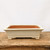 A rectangular, cream-colored glazed bonsai pot resting on a rustic wooden table against a plain white background. The pot has slightly flared sides, a rounded rim, and four small, decorative feet. While the exterior is finished with a smooth, off-white glaze featuring subtle speckling, the interior remains unglazed, showing its natural reddish-brown terracotta clay. The weathered wood of the table beneath displays a deep grain and rustic character, emphasizing the elegant simplicity of the ceramic bonsai container.