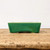 A long, descriptive alt text for the image is:

An eye-level, side-profile photograph of a shallow, rectangular bonsai pot featuring a vibrant emerald green glaze. The ceramic pot is characterized by an intricate and delicate crackle pattern (crazing) throughout the glaze, with deeper green tones pooling near the bottom edge. Its design includes slightly tapered sides and small integrated feet at the base for elevation and drainage. The pot sits on a rustic, weathered wooden plank, set against a clean, neutral white background that highlights the rich color and glazed texture of the piece.