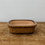 A shallow, rectangular ceramic bonsai pot with softly rounded corners is centered on a weathered, rustic wooden surface. The pot features a warm, mottled yellowish-brown glaze on its exterior, which has a subtle glossy sheen and dark brown highlights along the rim. The interior is unglazed, showing the natural, pale pinkish-tan color of the clay. The pot sits against a plain, light-gray background, which emphasizes its earthy tones and sturdy design. The lighting is soft and even, highlighting the textured grain of the wooden tabletop and the smooth, glazed surface of the bonsai container.