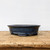 A sophisticated dark blue glazed oval bonsai pot sits centered on a rustic, weathered wooden surface. The pot features a deep navy glaze with a beautiful mottled or "oil spot" texture, giving it a rich and complex finish. Its elegant oval shape is complemented by a subtly flared rim and small, curved feet that elevate it slightly off the surface. The background is a clean, plain off-white wall, emphasizing the pot's deep colors and fine craftsmanship. This professional-grade ceramic container is designed to provide a striking and stable base for a wide variety of bonsai tree species.
