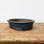 A front-facing, eye-level photograph of a shallow, oval-shaped glazed bonsai pot resting on a rustic wooden surface. The pot features a rich, dark blue glaze with a glossy finish and a complex, mottled texture. Small flecks of lighter cobalt and turquoise are visible throughout the deep navy glaze, creating a handcrafted appearance similar to a "hare’s fur" or oil-spot effect. 

The pot's design includes a wide, slightly flared rim and a subtle indentation around the base, just above its four small, rectangular feet. The interior of the vessel is left unglazed, showing the natural, warm reddish-brown color of the earthenware clay. It sits centrally on an aged wooden workbench with a prominent grain and several weathered marks, all set against a clean, neutral off-white background that highlights the pot's elegant silhouette and deep color.