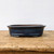A professional studio photograph of a shallow, oval-shaped dark blue glazed bonsai pot. The ceramic pot features a rich, glossy reactive glaze with deep navy tones and a subtle, speckled texture. It is designed with an elegant outward-curved rim and rests on small, integrated feet that provide slight elevation. The pot is positioned centrally on a rustic, weathered light-brown wooden tabletop, which shows a prominent natural wood grain. The background is a clean, solid white, creating a high-contrast setting that emphasizes the deep color and craftsmanship of the bonsai container.