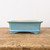 A side-profile view of a small, rectangular ceramic bonsai pot with a vibrant light blue glaze. The glaze features subtle texture and faint, darker speckling throughout. The pot’s design includes a slightly flared rim at the top and a matching decorative raised band along the bottom edge, resting on four small, curved feet that elevate the base. The pot is centered on a rustic, weathered wooden surface with a visible grain and aged texture, set against a clean, neutral white background that highlights the pot's soft color and simple silhouette.