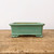 A side view of a rectangular, glazed green ceramic bonsai pot resting on a rustic, weathered wooden table. The pot features a soft celadon-green glaze with subtle variations and is designed with a flared upper rim and a distinct base ridge. It is supported by four small, decorative cloud-shaped feet that lift it off the surface. The interior of the pot shows a natural tan ceramic clay. The background is a clean, neutral white wall, highlighting the pot's elegant craftsmanship and soothing color.