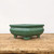 An eye-level studio photograph features a rectangular, green glazed bonsai pot resting on a rustic, weathered wooden tabletop. The ceramic pot, identified as model 1713, has a deep forest green glossy finish that reflects light along its smooth surfaces. Its design includes a thick, rolled-out rim and slightly outward-curving sides with subtle, wavy indentations for added texture. The pot is elevated by four sturdy, integrated feet with rounded "cloud" style edges. It sits centered against a minimalist, plain off-white background, highlighting the rich color and craftsmanship of the bonsai container.