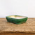 A rectangular, glazed ceramic bonsai pot in a deep, rich emerald green color sits centered on a weathered, rustic wooden surface against a plain off-white background. The pot features rounded corners and a slightly flared rim, supported by four small, integrated feet. The vibrant green glaze on the exterior has subtle, natural-looking variations in tone, while the interior is a contrasting pale cream color where the green glaze has softly bled over the top edge. This classic, high-quality bonsai pot provides an elegant and timeless vessel for miniature trees, highlighting the natural beauty of a bonsai display.