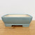 A side profile view of a 7-inch light blue glazed Yixing bonsai pot resting on a light-colored wooden table against a plain white background. This rectangular ceramic pot features a smooth, light blue glaze with delicate tonal shifts and rounded corners. It has a slight outward taper from its base to a thick, rounded rim. The pot sits on four small, rectangular feet, and its interior reveals a pale, off-white unglazed ceramic finish. This elegant and functional container is designed for small to medium-sized bonsai trees.