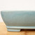 A side-profile close-up of a light blue glazed Yixing bonsai pot. The ceramic vessel features a soft, robin’s egg blue glaze with subtle mottling and a smooth, glossy finish that catches the light. This rectangular pot has gently rounded corners and a distinct stepped base that creates integrated feet, providing necessary elevation for drainage beneath the container. The pot is shown resting on a light-toned wooden surface against a clean white background, which emphasizes its elegant silhouette and the cool color tone of the glazed surface.