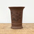 A tall, unglazed brown ceramic bonsai pot stands centered on a rustic, light-toned wooden surface against a plain, neutral white background. The pot features a cylindrical body that tapers slightly toward the bottom, ending in a sturdy pedestal base with a raised horizontal band for detail. Its wide rim flares elegantly outward at the top. 

The matte, chocolate-brown clay surface is decorated with a fine, black-etched illustration in a traditional style. The artwork depicts a lush peony bush in bloom, with two small birds perched on a branch to the left and a third bird in flight above the central flower. A small butterfly flutters near the upper right of the scene. To the left of the floral illustration, there is a column of vertical Chinese calligraphy, and a small rectangular seal mark is etched into the lower right side. The overall aesthetic is earthy and classical, highlighting the fine craftsmanship and textured finish of the unglazed clay.