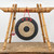 A mini brass gong hangs from a decorative Shao Shan bamboo stand, presented against a clean white background. The stand is crafted from thick, natural-toned bamboo poles, featuring a horizontal crossbeam supported by four angled legs that form a stable A-frame on each side. The bamboo joints are reinforced with neatly wrapped copper-colored wire, and small wooden pegs are integrated into the top bar.

Suspended from a bright red cord in the center of the stand is a circular brass gong. The gong's face is designed with three concentric circles: a wide, matte black outer ring, a textured, golden-tan middle ring with visible hammering marks, and a solid black center. Three red Chinese characters, “中国梦” (Zhōngguó mèng), meaning "Chinese Dream," are painted onto the top of the gong's outer ring and are mirrored on the center of the bamboo crossbeam above. 

To the left of the gong, a small wooden mallet hangs from a black cord. The mallet features a round, padded head with a classic red and white striped pattern. The entire ensemble sits on a rustic, weathered wooden surface, showcasing its artisanal quality as a decorative accessory often paired with bonsai trees or used in zen-inspired home decor.