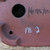 A detailed close-up shot of the underside of a reddish-brown, unglazed Bigei bonsai pot. The image shows a large, perfectly round drainage hole at the top left and a smaller wiring hole integrated into a rounded corner foot at the bottom left. To the right of the large hole, three Japanese characters, '美芸造' (Bigei Zou), are hand-incised into the clay, serving as the artist's signature and indicating it was made by the Bigei kiln in Tokoname, Japan. Below the signature, there are faint markings in white chalk, likely used for inventory purposes. The pot's surface has a natural, slightly textured finish typical of high-quality Tokoname clay.