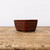 A small, rectangular unglazed Japanese bonsai pot by Bigei, known for Tokoname craftsmanship. The pot is made of deep reddish-brown clay and features a decorative relief of a basketweave pattern on its side panels, separated by smooth vertical bands. It has slightly flared sides and sits on small, rounded feet. The pot is centered on a rustic, weathered wooden table against a plain, off-white background, highlighting the intricate texture and handcrafted quality of the ceramic.