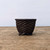 A miniature, round Japanese Tokoname bonsai pot, masterfully crafted by the renowned Bigei kiln, sits centered on a rustic, light-toned wooden surface. This small ceramic pot is characterized by its rich, matte chocolate-brown color and an intricate, hand-carved basket weave pattern that covers its entire body, mimicking the texture of a woven wicker basket. The top edge is finished with a thick, twisted rope-style rim, adding to its artisanal appeal. The pot is supported by three small, sturdy, integral feet in a tripod arrangement. The clean, off-white background emphasizes the detailed texture and earthy aesthetic of this high-quality unglazed ceramic piece, making it ideal for a small or shohin-sized bonsai tree.
