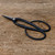 A pair of professional Kaneshin 36B bonsai shears rests on a rustic, light-brown wooden surface. These authentic Japanese bonsai tools feature a sleek, matte black finish on the body and handles. The sharp, pointed blades show a sliver of polished steel along the cutting edge, held together by a prominent silver, textured pivot screw. Gold-colored Japanese kanji characters are delicately engraved near the joint. The long handles curve outwards into two large, ergonomic oval finger loops, designed for precision and comfort during bonsai pruning and maintenance. The weathered wooden background, marked with faint scratches and grain patterns, provides a natural contrast to the refined, dark silhouette of the shears.