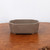 A high-quality, unglazed bonsai pot made of earthy brown clay, featuring a rectangular shape with distinctively notched or bracketed corners. The pot has a smooth, matte finish and sits on subtle integrated feet that provide stability and allow for proper drainage. It is positioned centrally on a polished, warm-toned wooden surface with a rich grain pattern, set against a plain off-white background. This professional photograph showcases the pot's minimalist design and natural texture, characteristic of traditional unglazed ceramic bonsai containers often used for coniferous or deciduous trees to create a grounded, natural aesthetic.