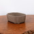 An unglazed brown ceramic bonsai pot, model 1509, is showcased on a polished wooden surface. The pot features a rectangular form with subtle, rounded lobes at each corner, giving it an elegant, artisanal appearance. It sits on four small, integrated feet that provide a slight lift for drainage and aesthetics. The material has a smooth, matte finish in a warm, earthy brown clay tone, typical of traditional unglazed bonsai containers. The pot is centered in the frame against a minimalist off-white background, resting on a richly grained, warm-toned wood tabletop that highlights the pot's clean lines and natural texture.