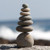 A septuple rock cairn composed of seven smooth, rounded river stones meticulously balanced in a vertical stack. The stones, ranging in size from a large, sturdy base to a small, circular top piece, sit atop a weathered gray rock. The composition is set against a soft, out-of-focus backdrop of a coastline, where the deep blue of the ocean meets the white froth of crashing waves. Natural sunlight illuminates the stones from the side, casting gentle shadows that define their organic shapes and earthy gray and tan tones. This tranquil arrangement of balanced stones evokes a sense of peace and mindfulness, serving as a perfect example of natural art for a Zen garden or as a complementary accessory for a bonsai display.