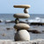 A serene medium-sized rock cairn stands perfectly balanced against the backdrop of a blurred coastal scene. The stack consists of six smooth, light-colored river stones arranged in an intricate "side2side" or zig-zag pattern. It begins with a wide, dome-shaped base stone that supports a flat, oval rock. Above this, the weight shifts laterally using smaller rounded stones as pivot points for alternating long, flat rocks that extend to the left and right. 

The stones' natural textures of grey and tan are highlighted by bright daylight. In the background, the soft focus reveals a deep blue ocean with white foamy waves and a clear sky, emphasizing the cairn's peaceful and meditative presence. This artistic arrangement of stacked rocks serves as a symbol of stability and zen-like balance, often used as a decorative accent in outdoor gardens or alongside bonsai trees.