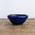 A detailed view of a handcrafted blue glazed ceramic bonsai or succulent pot. The small bowl features a deep cobalt blue glaze with a glossy finish that shows subtle color variations and a delicate crackle texture. Its organic, hand-pinched form is characterized by an irregular, slightly wavy rim and a tapering body that rests on a visible, unglazed brown clay foot. The pot is positioned on a rustic, weathered wooden tabletop set against a plain, light-colored background, highlighting its artisanal quality and vibrant color.
