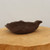 A side-view photograph of a dark, chocolate-brown unglazed Japanese bonsai planter. This artisanal ceramic pot has a hand-molded, organic form with an uneven, wavy rim and a textured, matte finish, giving it a rustic, natural appearance. It rests on a light-colored, circular wooden slab against a clean, off-white background, highlighting its unique silhouette and earthy texture.