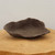 A handcrafted, unglazed ceramic bonsai pot in a dark, earthy charcoal brown. This unique Japanese-style planter features a shallow, irregular shape with organic, undulating edges that resemble a piece of naturally formed stone or a large seashell. The entire surface has a rustic, tactile texture, showcasing its handmade quality. Two small, circular drainage holes are present in the center of the pot's interior. The pot sits on a polished, light-colored wooden platform, set against a clean, neutral off-white background. Its wabi-sabi aesthetic makes it an ideal choice for a naturalistic bonsai composition.