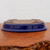A detailed, upside-down view of a shallow, oval Japanese bonsai pot, highlighting its construction and finish. The exterior features a rich, glossy cobalt blue glaze that catches light and reflections from the surroundings. The unglazed bottom reveals a matte, reddish-brown terracotta clay, featuring two large, circular drainage holes and four subtle, integral feet that provide a small amount of lift. The pot rests on a warm-toned, polished wooden surface with a decorative, rugged natural edge, set against a neutral, off-white background.