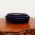A set of three nested, oval-shaped Japanese bonsai pots featuring a deep, glossy cobalt blue glaze. The ceramic pots are stacked from largest to smallest on a polished, reddish-brown wooden display slab with a rustic, textured edge. Each pot has a smooth, rounded rim and a high-shine finish that reflects the light. The vibrant blue of the glazed stoneware stands out against a clean, neutral off-white background, highlighting the classic design and elegant craftsmanship of these traditional bonsai containers.
