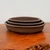 An professional studio photograph shows a set of three oval-shaped, unglazed bonsai pots nested within one another, showcasing their graduated sizes. Made from dark chocolate-brown clay with a smooth, matte finish, these Japanese-style planters feature a minimalist design with straight sides and a narrow, flat rim. The pots are elegantly displayed on a polished, warm-toned wooden surface with a visible natural grain and a rustic, irregular edge in the bottom right corner. The neutral, off-white background creates a clean contrast, highlighting the rich, earthy tones and professional craftsmanship of the ceramic containers, which are ideal for traditional bonsai cultivation.