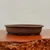 A side-profile shot of a shallow, oval-shaped bonsai pot made from unglazed, dark brown ceramic. The planter has a smooth, matte finish and a flat rim that curves slightly outwards. It is elevated by small, low-profile feet. The pot sits on a polished reddish-brown wooden stand with a heavily textured, natural live edge in the foreground. The background is a plain, light-colored wall, emphasizing the simple and elegant design of the traditional Japanese-style bonsai planter.