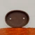 A high-angle, top-down view of an oval, unglazed brown ceramic bonsai pot. The shallow planter is a deep, matte chocolate brown and features two circular drainage holes at its base for optimal water drainage and root health. It is positioned against a plain, light-gray wall and sits behind a textured, vibrant orange-brown wooden bark surface in the foreground. This minimalist Japanese-style pot is designed for cultivating and displaying bonsai trees, emphasizing natural earthy tones and a clean, functional design.