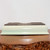 A rectangular cream Japanese glazed bonsai pot is shown upside down on a polished, dark brown wooden surface. The pot features a smooth, glossy pale cream glaze with subtle hints of light green that wraps around its sides and slightly flared top rim. The unglazed base, facing upward in this perspective, reveals a dark grey-brown clay material and highlights four wide, low-profile feet designed for stability and drainage. The pot's design is elegant and minimalist, with soft rounded corners and a subtle rectangular indentation along its side. This Japanese ceramic bonsai pot from Bonsai Outlet is photographed against a plain light grey background, emphasizing its clean lines and soft color palette, which would complement a wide variety of bonsai tree species and styles.