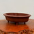 A horizontal photograph of a traditional Yixing unglazed bonsai pot, crafted from classic reddish-brown purple clay. The vessel features a distinctive mokkou (lobed flower) shape with four gentle curves along its wide rim and body, creating a soft, cloud-like silhouette. Its unglazed surface has a smooth, matte finish that highlights the natural beauty of the clay. The pot is elevated by four small, rounded, stubby feet for drainage and aesthetic balance. It sits atop a highly textured, polished wood burl display stand with a rugged, irregular natural edge. The entire scene is set against a minimalist, neutral off-white background, emphasizing the artisanal craftsmanship and elegant form of the bonsai pot.
