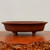 A side-profile photograph of a traditional Yixing unglazed bonsai pot, showcased on a polished, live-edge wood display stand. The pot is shallow and oval with a smooth, matte finish in a rich reddish-brown terra cotta color, characteristic of high-quality Yixing clay. It features a rounded, slightly flared top rim and is supported by four subtle, tapered feet. The clean, minimalist design is set against a neutral, off-white background, highlighting the pot's elegant proportions and artisanal craftsmanship.