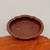 A top-down view of a professional-grade, unglazed Yixing bonsai pot in a deep reddish-brown clay. The pot features a distinctive scalloped or lobed oval rim, creating an elegant cloud-like silhouette. Inside the smooth, matte interior, two large circular drainage holes and four smaller wire holes are visible at the bottom, essential for healthy tree growth and stability. The pot is displayed on a textured, warm-toned wooden surface against a neutral, off-white background, emphasizing its classic aesthetic and functional design for bonsai enthusiasts.