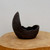 A side-profile photograph of a small, unglazed ceramic succulent planter with a striking, organic shape. The dark brown, almost black, pot is asymmetrical, resembling a cracked eggshell or a crescent moon with one side sweeping up into a tall, graceful point and the other forming a shallow, rounded bowl. Its surface has a rough, matte, and weathered texture with visible pits and an uneven, jagged rim that emphasizes its rustic, handcrafted quality. The planter is centered on a light-colored wooden slab with a natural grain, set against a plain, neutral background. This artistic container is a characteristic example of an unglazed bonsai pot, perfect for showcasing a single succulent or a small bonsai tree in a naturalistic style.