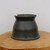 A professional studio photograph of a dark charcoal gray ceramic bonsai pot, handcrafted by American potter Joshua Allen Silvia. The pot features a unique, modern conical shape with a wide, flared base and a slightly flared top rim that includes two square-cut notches on opposite sides. The stoneware has a semi-matte finish, with a smooth upper half and a rugged, horizontally textured band around the lower section, giving it an organic, earth-toned appearance. The pot rests on a rustic, cross-cut natural wood slab, set against a clean, neutral white background to highlight the craftsmanship and dark glaze of this handmade American bonsai container.