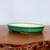 A shallow, oval-shaped glazed bonsai pot is showcased in a side-profile view, resting on a polished, dark wood surface. The pot features a vibrant, emerald-green glaze on its exterior, characterized by a fine, intricate crackle pattern, also known as crazing, which adds a textured, vintage appeal. 

The top edge has a smooth, rolled rim that curves slightly outward. Inside, the pot is a contrasting smooth, off-white or cream color. The vessel is supported by small, inconspicuous feet at the base, elevating it slightly off the wooden table. The background is a clean, neutral off-white wall, ensuring the focus remains entirely on the elegant craftsmanship of the ceramic bonsai container.