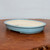 An eye-level, slightly angled photograph of a shallow, oval-shaped ceramic bonsai pot. The exterior of the pot is finished in a smooth, glossy, light sky-blue glaze, while the interior remains an unglazed, matte cream color. The pot features a rounded rim and four small, integrated feet that provide a slight elevation. A single drainage hole is partially visible at the bottom of the basin. The pot sits on a dark, rustic wooden surface with a prominent grain, set against a plain, light grey background, highlighting the pot's elegant and minimalist design.