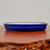 An eye-level, close-up photograph showcases a shallow, oval-shaped bonsai pot with a vibrant, high-gloss cobalt blue glaze. The pot features a smooth, rounded rim and a low profile, resting on a polished, rich brown wooden surface that reflects its base. The interior of the container is left unglazed, showing the natural, light-beige color of the ceramic material. The background is a minimalist, soft-grey wall, which emphasizes the deep blue hue and clean lines of the pottery. This elegant glazed ceramic pot, designed for both aesthetic appeal and practical bonsai cultivation, is a representative example of the high-quality specialty containers available at Bonsai Outlet.