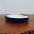 A high-quality 7-inch oval glazed bonsai pot with a deep cobalt blue exterior and a contrasting creamy-white interior. The ceramic pot features a smooth, rolled rim and a shallow profile specifically designed for traditional bonsai tree cultivation. It is positioned on a rustic dark wood surface against a plain, neutral light gray background. A drainage hole is partially visible at the center of the interior base, essential for proper soil aeration and moisture management. The glossy finish of the glaze highlights the rich blue pigment, making it an elegant container choice for various bonsai styles and species.