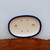 A top-down view of an oval-shaped glazed ceramic bonsai pot, showcasing its deep cobalt blue rim and smooth, cream-colored interior. The pot is designed for functionality and health, featuring six pre-drilled holes at the bottom: two large circular drainage holes located on the left and right sides, and four smaller wiring holes positioned in the center to help secure a tree firmly in place. The pot rests on a rustic, reddish-brown wooden slab with a natural, textured edge, set against a plain, light grey background that highlights the pot's clean lines and vibrant glaze.