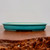 A side-profile view of a shallow, oval-shaped glazed bonsai pot, showcasing its vibrant turquoise or teal color with a subtle, fine crackle pattern across the exterior. This low-profile ceramic container features a slightly flared rim and is elevated by small, integrated feet at its base to allow for drainage. The pot sits atop a smooth, polished wooden table with a rich brown finish, while a textured, rustic wooden edge is partially visible in the bottom foreground. The interior of the pot is a contrasting unglazed creamy white. The clean, neutral background emphasizes the pot's elegant form and striking glaze, illustrating its suitability for various bonsai tree styles and displays.