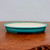 A shallow, oval-shaped glazed bonsai pot in a vibrant teal-blue hue sits centered on a warm, reddish-brown wooden surface. The exterior of the pot features a fine crackle glaze, also known as crazing, which creates a delicate network of lines across the turquoise finish. Along the thin, slightly outward-turned rim, subtle dark speckles are visible. The interior of the pot contrasts with the exterior, showing a smooth, off-white or cream-colored glaze. The pot is elevated by small, integrated feet at its base, designed for drainage and airflow in bonsai cultivation. It is photographed against a neutral, plain light-grey background, which emphasizes the pot's elegant form and textured finish.